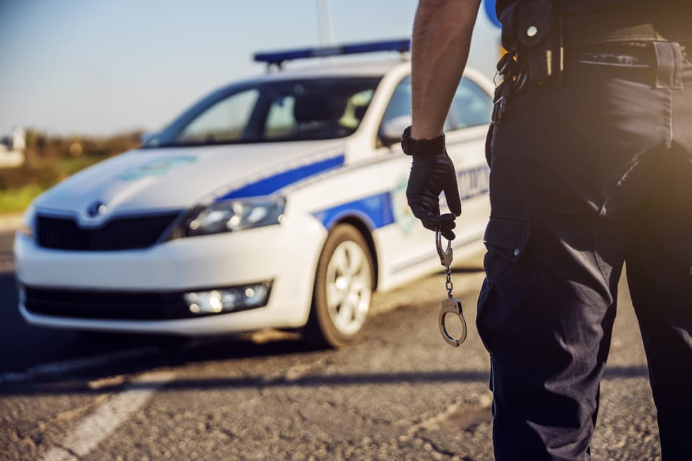 Police officer holding handcuffs, police car in background.